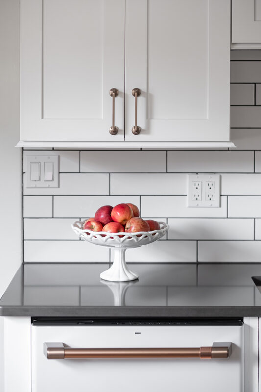 Shows dishwasher with gold handle, black cabinets, and white subway tile backsplash. The backsplash has black grout