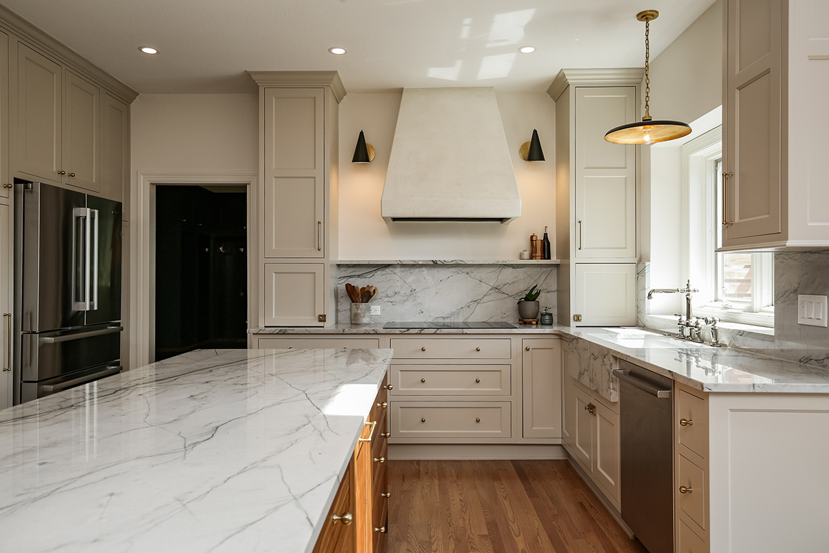 marbled white stone island countertop sits on bright wooden cabinetry with brass accents. Marbled white stone countertops sit on the white kitchen cabinetry. Marbled stone backsplash.