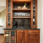 mini bar with brown wooden cabinets. The minifridge has protein drinks and carbonated water inside. The wine cooler has various bottles of wine. There is glassware filling the shelves.