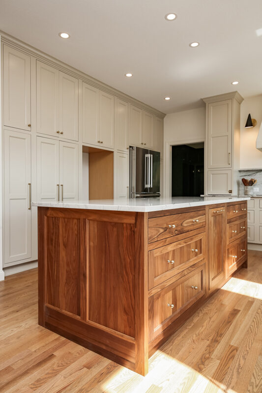 brown wood kitchen island with copper knobs sits beneath a marbled white countertop. The rest of the kitchen has modern appliances and white cabinetry.