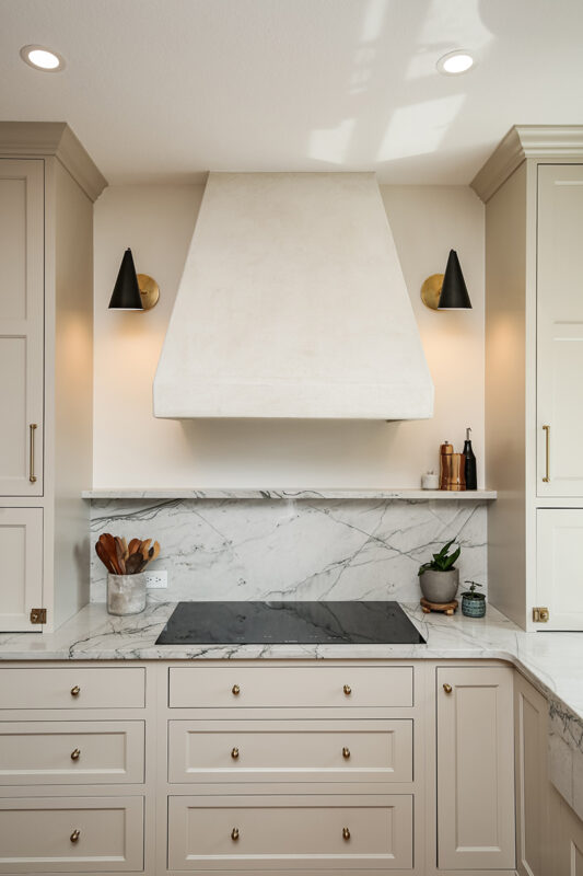 Closeup shot of stovetop and cabinetry. The cabinetry is all white with brass knobs. The range hood is also white. The stovetop is electrical and modern. Marbled white countertops.
