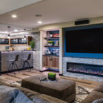 wide view of the basement with wet bar and large screen tv. below the tv is a built in fireplace.
