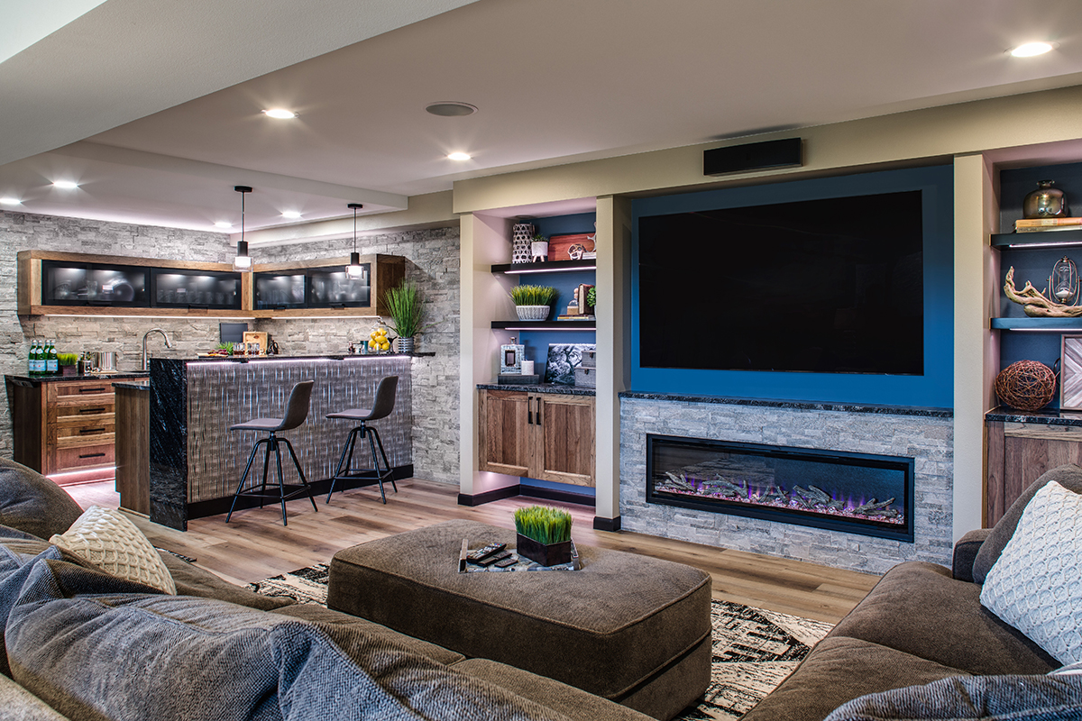 wide view of the basement with wet bar and large screen tv. below the tv is a built in fireplace.