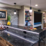 The countertop of the bar. Dark grey countertop paired with brown wood cabinets, and a tiled featured wall.
