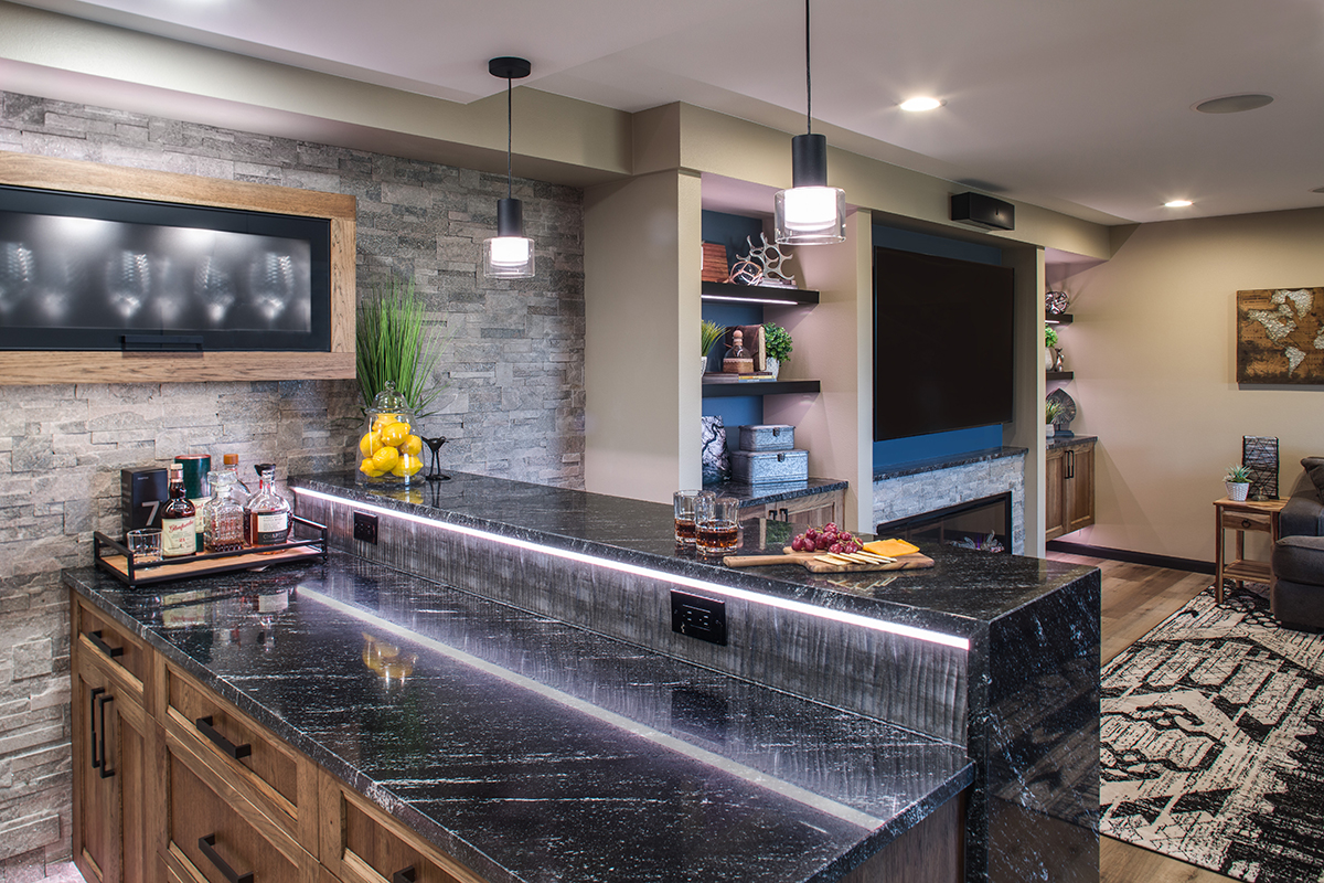 The countertop of the bar. Dark grey countertop paired with brown wood cabinets, and a tiled featured wall.
