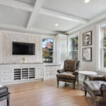 Shows a peak of the piano on the left side of the photo with sitting room on the right side of the view. There are two overstuffed chairs that line the left wall. The built in cabinets are along the back wall.