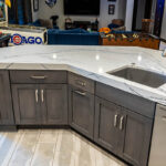 Shows the back side of the wet bar. There are grey cabinets under the sink with stainless steel hardware. The counter top has a sink and the countertop is white with a swirled pattern.