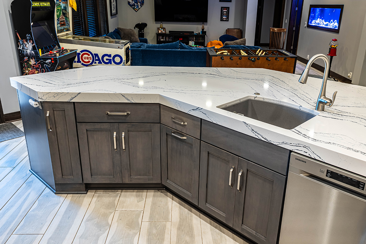 Shows the back side of the wet bar. There are grey cabinets under the sink with stainless steel hardware. The counter top has a sink and the countertop is white with a swirled pattern.