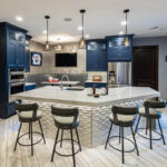 Another view of the kitchen that shows the booth, bar top, stainless steel built-in ovens, double fridge. The cabinets are dark blue and the island is unique.