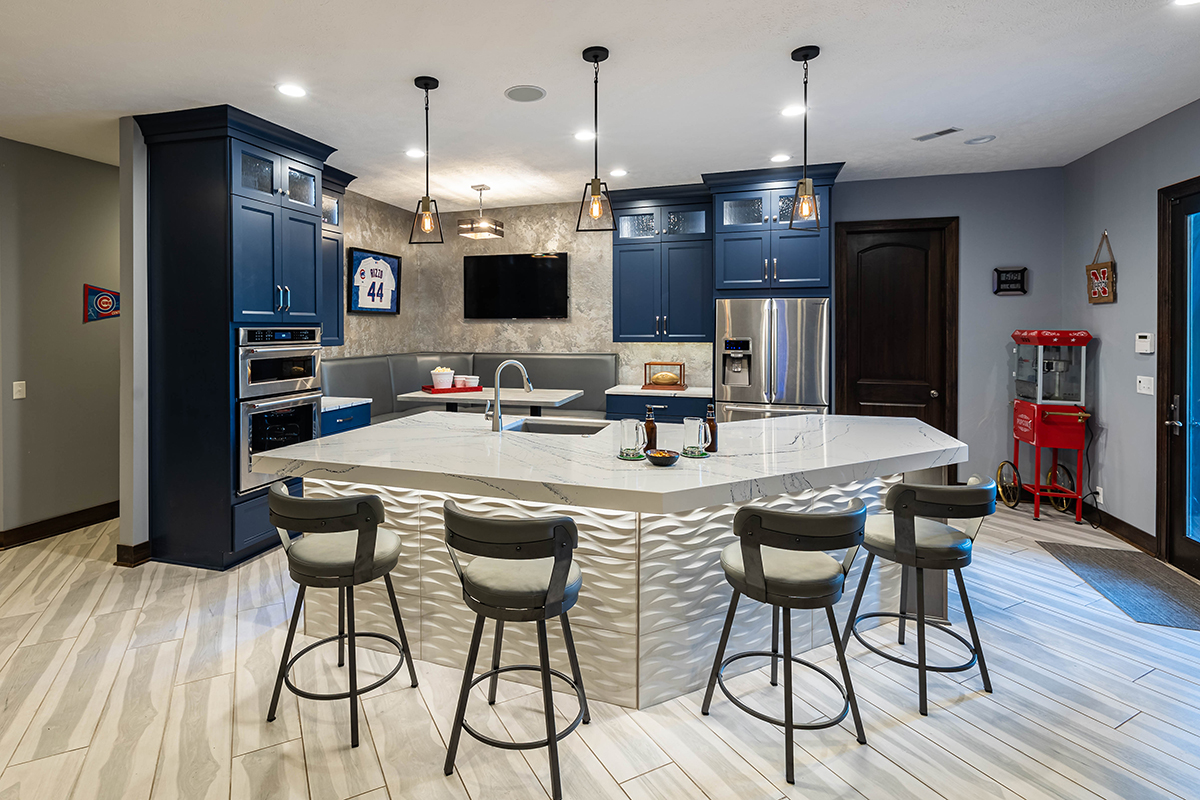 Another view of the kitchen that shows the booth, bar top, stainless steel built-in ovens, double fridge. The cabinets are dark blue and the island is unique.