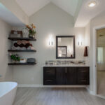A large soaking tub is off the left of the picture. There is a floating vanity featured with dark brown wood, silver hardware, a tile backsplash that matches the shower. There are three floating shelves that have extra towels and decorations.
