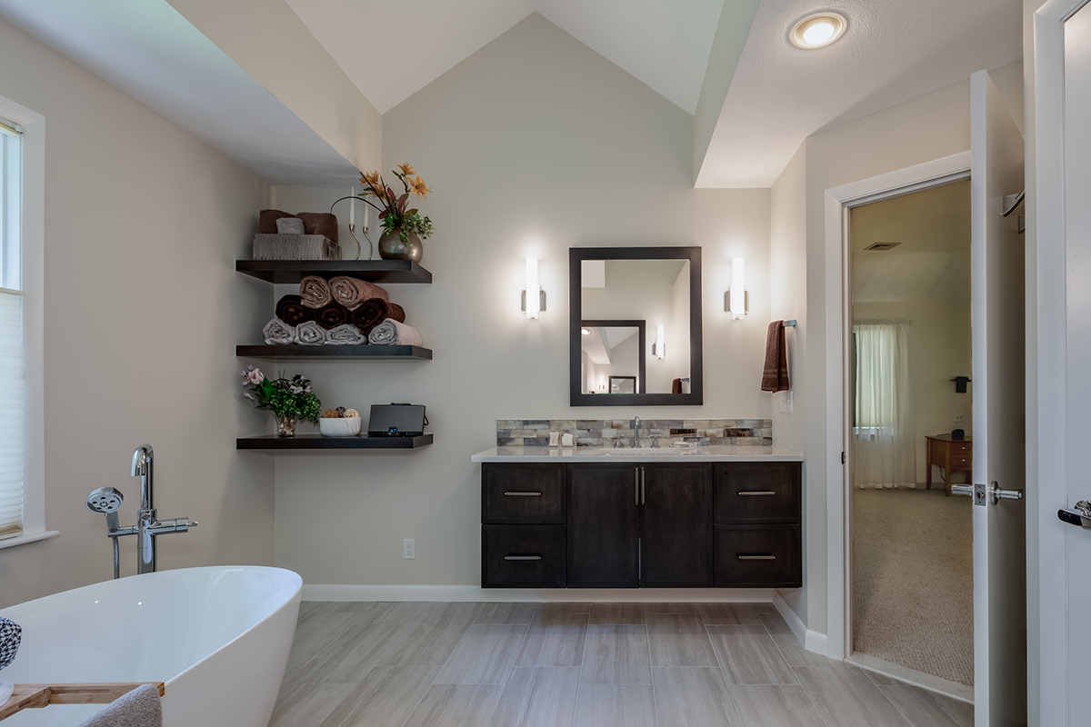 A large soaking tub is off the left of the picture. There is a floating vanity featured with dark brown wood, silver hardware, a tile backsplash that matches the shower. There are three floating shelves that have extra towels and decorations.