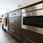 Shows the large island with dark cabinets and silver handles. There is a built in stove and microwave in the base of the island.
