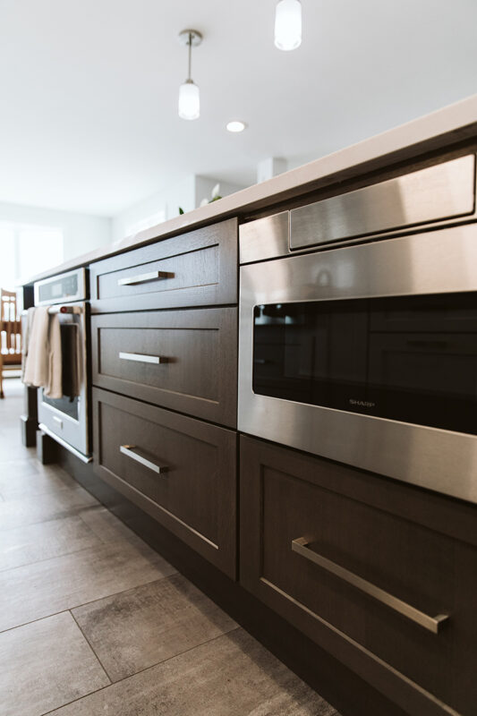 Shows the large island with dark cabinets and silver handles. There is a built in stove and microwave in the base of the island.