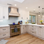 Brushed light brown cabinets are on the lower half of this kitchen with crisp white cabinets on the top. The counter tops are white and there is a wavy light blue backsplash.