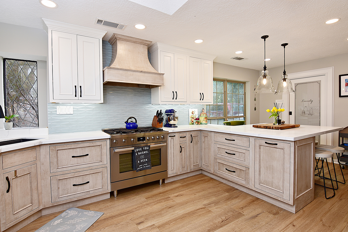 Brushed light brown cabinets are on the lower half of this kitchen with crisp white cabinets on the top. The counter tops are white and there is a wavy light blue backsplash.