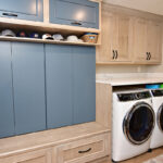Built-in locker style storage in half of the room with the washer and dryer on the other side of the room. There are blue accents on the back wall of the locker and the upper cabinet faces.