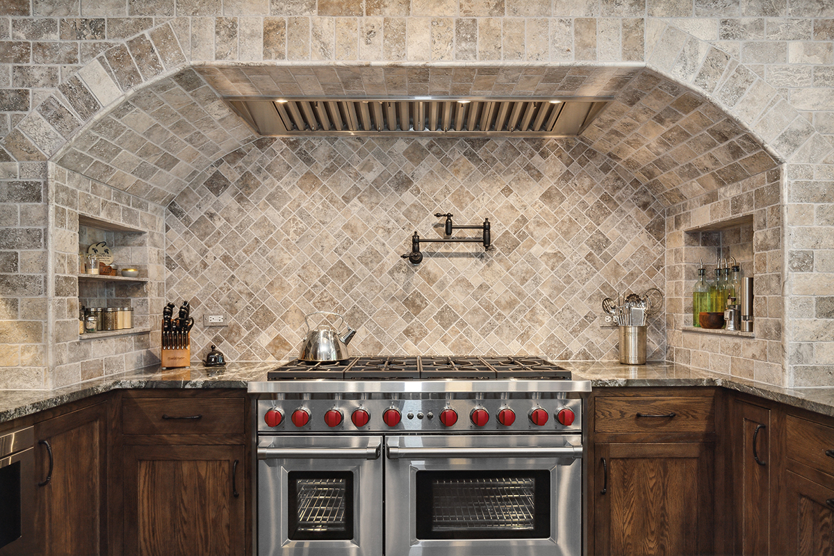 large stone exhaust hood with stone backsplash and wall pattern surrounding brown kitchen cabinetry and stainless steel stove.
