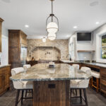 brown kitchen cabinetry with stone backsplash, floors, and countertops. White cushioned seats are tucked under the large island.