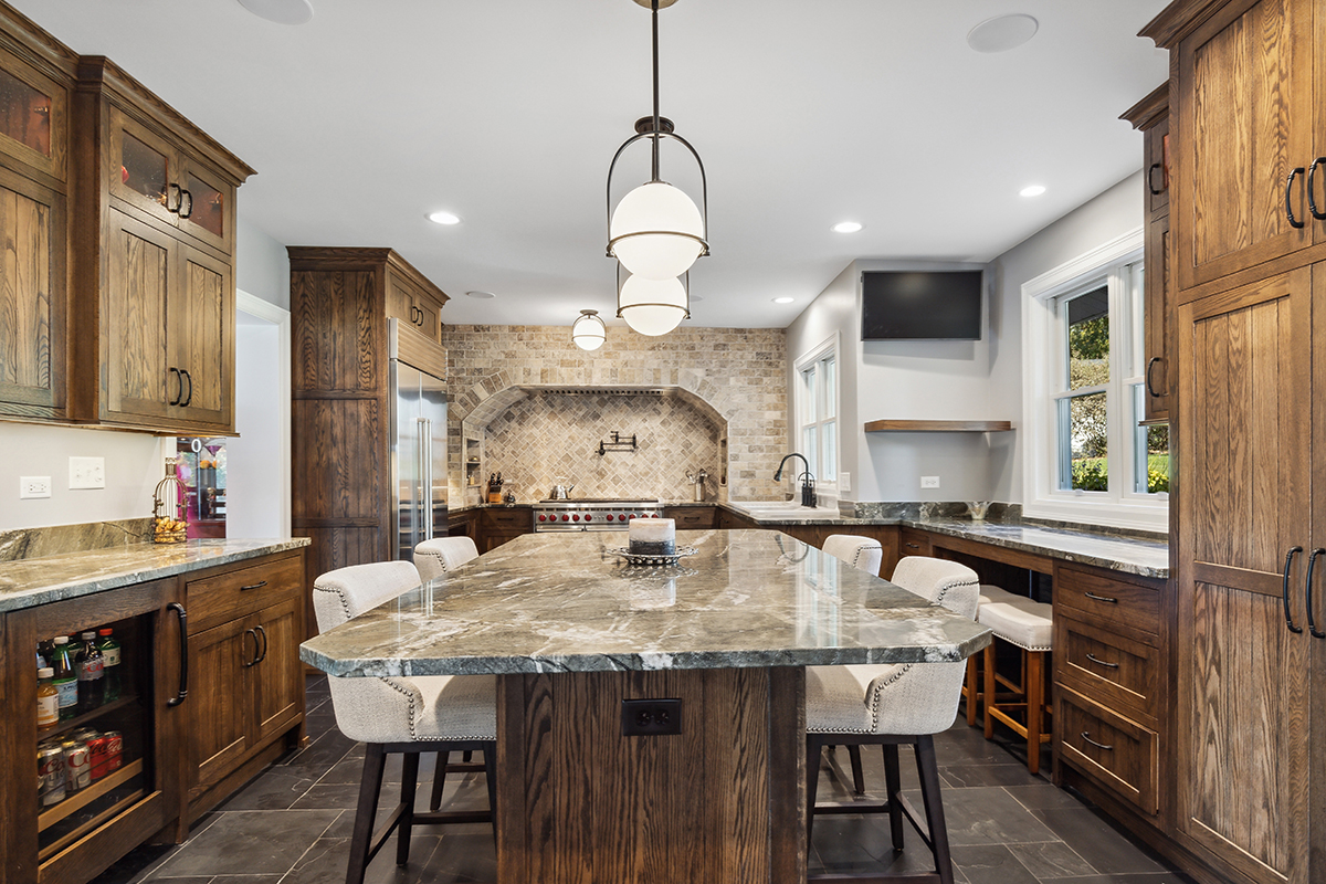 brown kitchen cabinetry with stone backsplash, floors, and countertops. White cushioned seats are tucked under the large island.