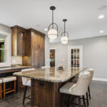 dark wooden cabinetry in kitchen. Stone floors and stone countertops. White cushioned stools are tucked under the island.