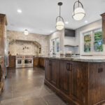 large kitchen with wooden grainy cabinetry. Large range hood with intricate stone backsplash sits above stovetop.