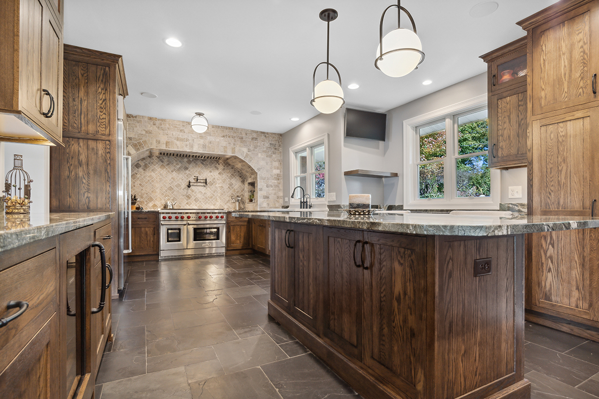 large kitchen with wooden grainy cabinetry. Large range hood with intricate stone backsplash sits above stovetop.