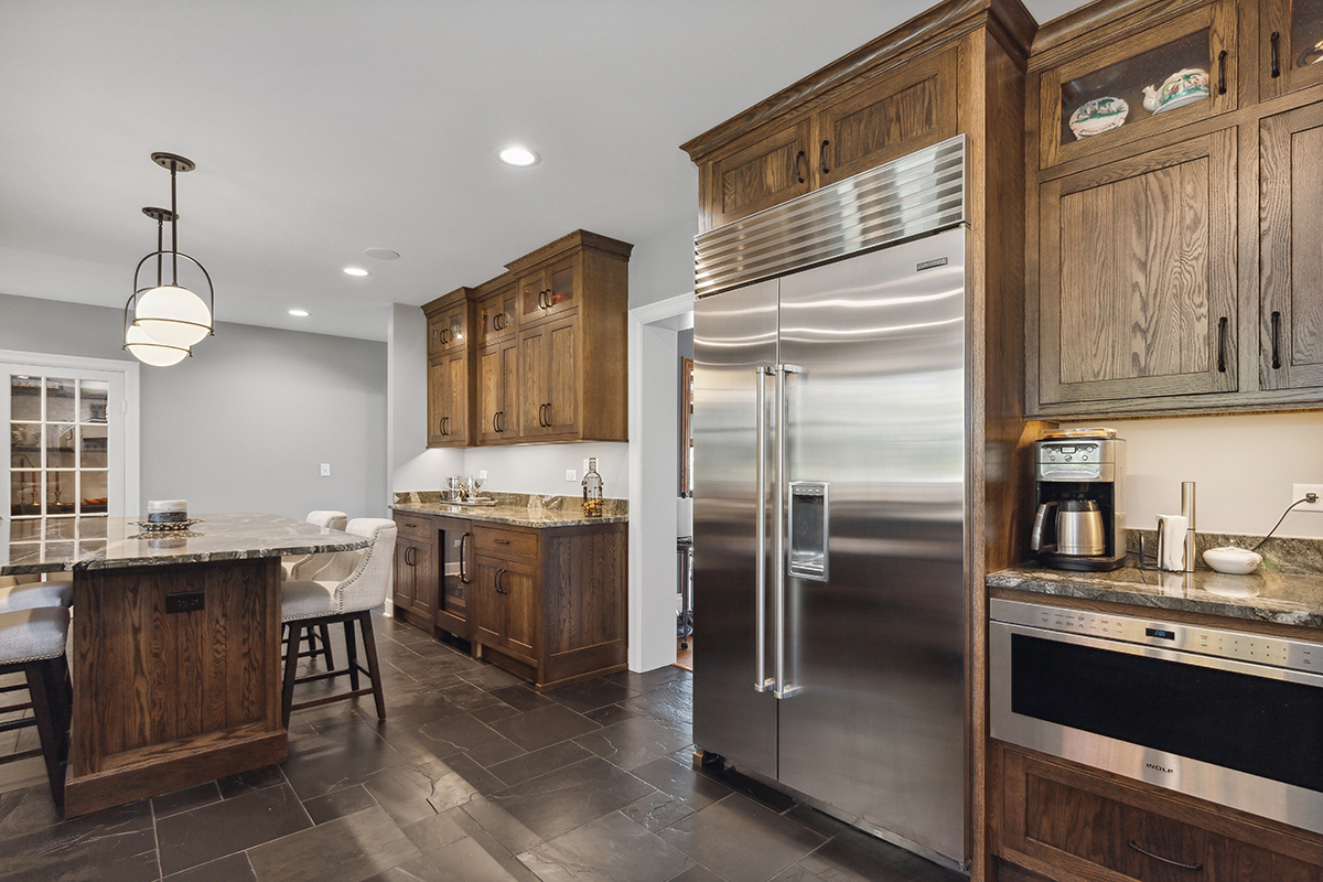 large stainless steel fridge inside dark wooden cabinetry. open kitchen with stone countertop island.