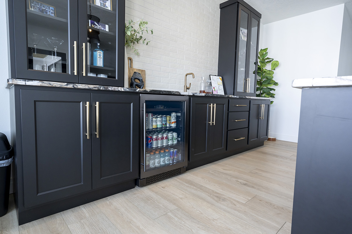 black minibar with marbled stone countertop. The cabinets are filled with beverages and glassware, and sit next to a white brick backsplash.