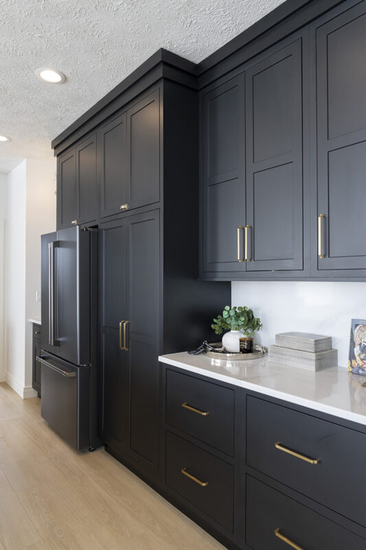 This kitchen has white counter tops with black cabinets low. In this picture we see a large pantry and the fridge. The black cabinets go from floor to ceiling in this photo.
