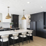 dark black kitchen cabinetry with gold accents. White island stone countertop with white stools.