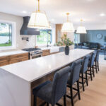 Open kitchen with long island with seating facing stained base cabinets, oven, hood, sink and large windows overlooking a lake.