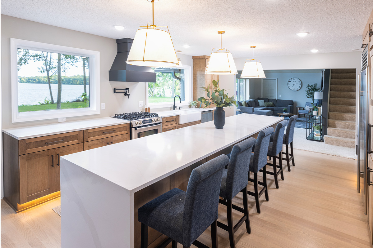 Open kitchen with long island with seating facing stained base cabinets, oven, hood, sink and large windows overlooking a lake.