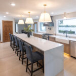 Open kitchen with long island with seating facing stained base cabinets, oven, hood, sink and large windows overlooking a lake.