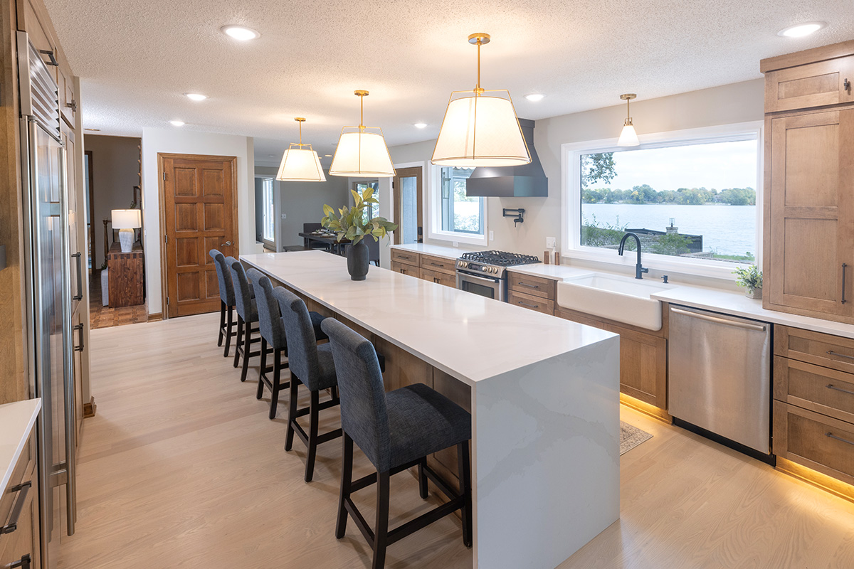 Open kitchen with long island with seating facing stained base cabinets, oven, hood, sink and large windows overlooking a lake.