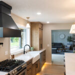 Open kitchen with long island with seating facing stained base cabinets, oven, hood, sink and large windows overlooking a lake.