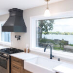 Open kitchen with long island with seating facing stained base cabinets, oven, hood, sink and large windows overlooking a lake.