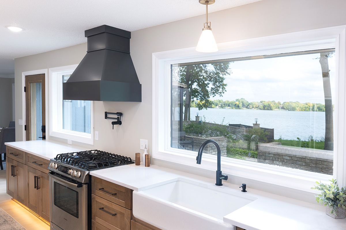 Open kitchen with long island with seating facing stained base cabinets, oven, hood, sink and large windows overlooking a lake.