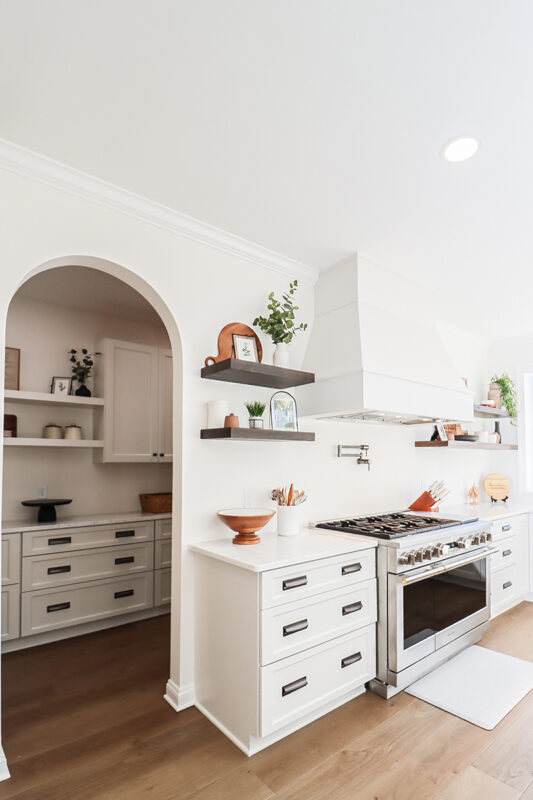 White kitchen cabinets with large hood. View looks into a pantry in arched walkway.