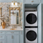 Light blue bathroom vanity cabinets with ornate gold mirrors and sink hardware. Showing open cabinet with stacked washer and dryer.