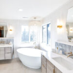 Large master bathroom with 2 vanity areas with light Montana stain, powder desk and large free-standing tub in the corner below large windows.