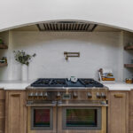 Light brown-stained lower and kitchen cabinets with gray painted archway blower above a professional range.