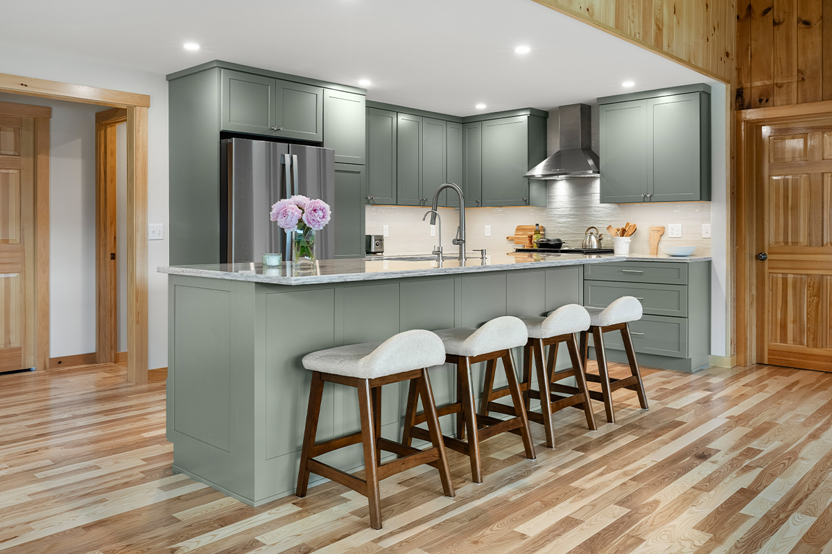 Green kitchen cabinets with island seating and light brown wood floors.