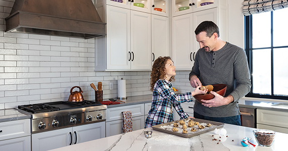 A husband and wife smiling in their kitchen that has white inset cabinets, white subway tile backsplash, and white marble-style countertops.